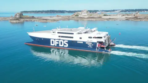 A DFDS boat heads out of a harbour in Jersey. The vessel is blue and white with the ferry firm's logo on its side in large white block capitals. It is sailing past Elizabeth Castle.