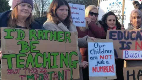 Staff hold handmade placards that read 'I'd rather be teaching but this is important' and 'Our children are not just a number'