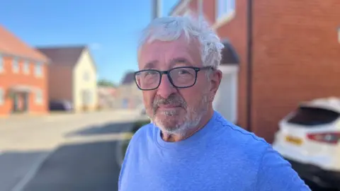 Henry Godfrey-Evans/BBC A man with glasses and a blue T-shirt standing in the middle of the street, which has modern looking red-brick homes on it.