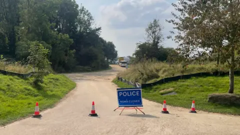 A blue police sign and orange cones blocking the gravel path up to Ham Hill. Either side of the path is surrounded by trees and grass.