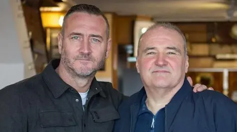 Actor Will Mellor, with his arm around the shoulder of Lee Castleton. Mellor has cropped dark hair and a short beard and is wearing a black jacket. Mr Castleton is waring a blue jacket and blue zip-up top.