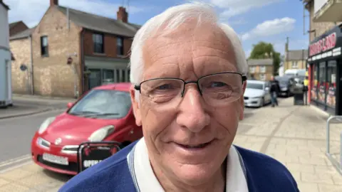 Michael Gibbs looking into the camera while standing in Whittlesey High Street. He has short white hair and glasses and is wearing a blue jumper.