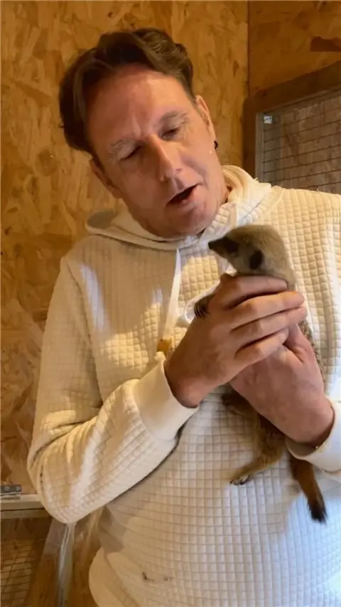 A man in a white jumper holds a meerkat.