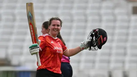 Kathryn Bryce raises her bat as she celebrates her century