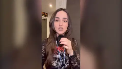 Woman with long dark brown hair holding a small microphone, looking straight at the camera