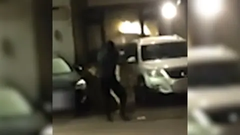 A man dressed in black firing a gun in a dark street in front of a house with a silver SUV.