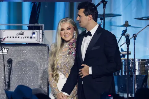 Charlie Kirk de Getty Images tiene la mano de Erikarene Franzbe en el escenario al lado del kit de batería y grandes altavoces plateados. Ella usa un vestido de astillas brillante y tiene el pelo largo y rubio. 
