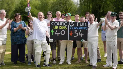 PA Media Sir Ed Davey pictured with a group of councillors who are wearing cricket gear. Sir Ed is holding a cricket bat up in the air, while holding a score board in the other which shows the Lib Dem's have gained 40 seats and the Conservatives have lost 34.