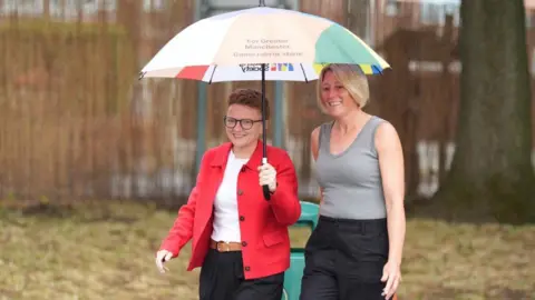 PA Media Labour's Bev Craig arrives at Broad Oak Primary in Didsbury with her partner Tracy to vote in the Greater Manchester mayoral by-election. She holding a multicolour umbrella as she walks towards the polling station.