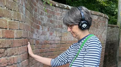 Karen Goddard Mrs Goddard is pictured resting her palm on a brick wall during the production of an audio trail she previously worked on. She has a pair of headphones on her head, covering her ears. She wears a white and blue striped long-sleeved top with a green bag strap over her torso.
