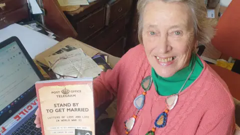 Anne Holland Anne holding her book up to the camera and smiling. She is wearing a green t-shirt a pink jumper and a colourful, chunky, acrylic necklace.