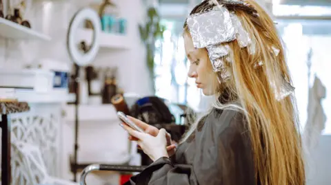 A blonde woman with foil in her hair as she gets her hair highlighted at a hairdressers. She is sat on a chair looking at her phone.