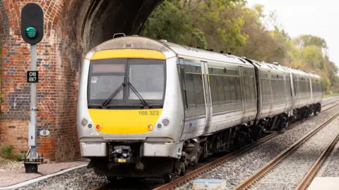 An East West Rail test train on railway tracks under a railway bridge, with trees lined beside the tracks