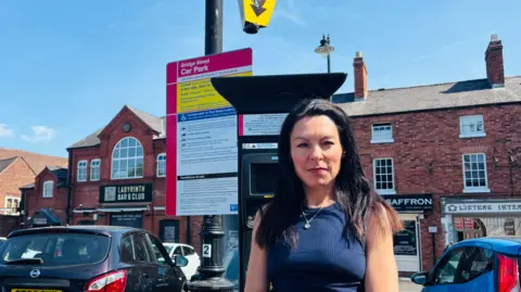 A woman with long black hair, wearing a blue vest top, stands by a parking machine in a town centre car park in front of a row of brick-built shops and restaurants 