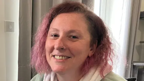 A woman with curly pink hair and a black nose ring smiles at the camera.