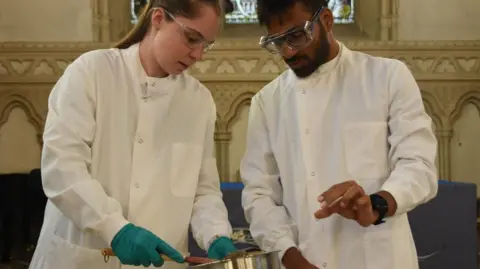 Jennifer Palmer with hair pulled back wearing blue gloves and Satish Viswanathan, with dark hair and beard, both wearing white lab coats and looking down at bowl