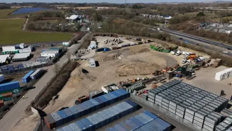 A photo taken from a drone, showing a stretch of industrial land with cars and lorries parked on it. There's a road on the left hand side and a motorway on the right.