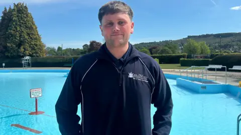 Jessica Bayley/BBC A young man wearing a navy uniform with a polo T shirt and fleece stands in front of a lido.