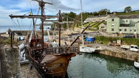 A view of Charlestown harbour. It has two stone walls forming a pincer. There is a small white watchtower building at the far end on the left. On the left in the foreground is a moored old-style wooden sailing ship. A more modern yacht is ashore on the right. On the upper right of the picture is the Pier House pub, painted in a matt light pea green.