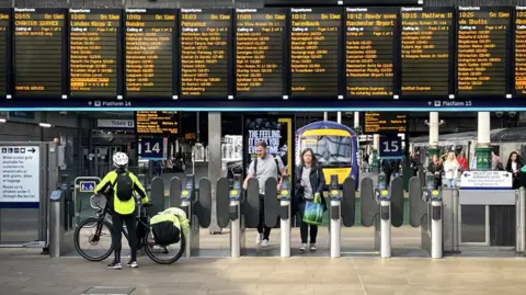 PA Media Train station platform gates with cyclist with bike in front looking up at departure board. couple walk through gates at other side with train in background.