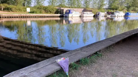 Luke Deal/BBC A bouquet of flowers left next by the side of the water, which has a row of five white and blue boats moored in it.