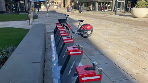 One bike and seven docks, six of which are empty. They are grey and red. It is places on a pedestrianised street.