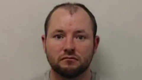 A poorly lit, grainy police mugshot of James Thomson, who has short dark hair and a beard. He is staring expressionless at the camera. Thomson is wearing a grey t-shirt and has been photographed against a grey background, without the use of a flash.