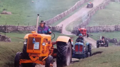 Dave Harrison Dave Harrison riding a yellow tractor on a green field, several over tractor trail behind him, as the group follow a countryside route in convoy.