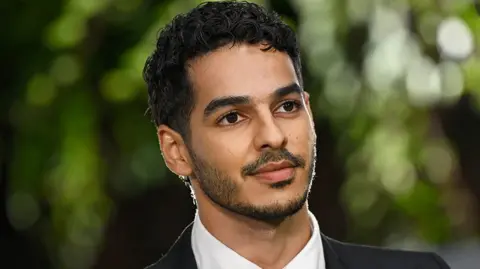 Getty Images Ishaan Khatter, um ator masculino em uma estreia olhando para longe, vestindo uma camisa branca e um blazer preto. O fundo é de árvores verdes borradas e fora de foco.