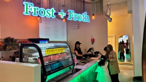 Part of the foodhall. We can see a business called Frost + Froth selling lots of different coloured drinks. A woman in a black top and a hat is serving a family. There is a woman wearing a hijab and two children with her. She is paying for her items.