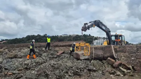 Government of Jersey A group of workers and a crane working on a damaged seawall. The crane is lifting a large piece of stone wall with the workers helping with the works. In front of the wall is rocks covered in seaweed and shells. 