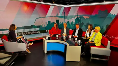A female presenter in an armchair in a TV studio faces 5 candidates on a red sofa. Four are women with one man in the middle