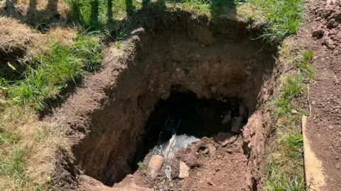 The image depicts a large hole dug into the ground, exposing a section of underground piping. Part of the piping remains exposed as brown sewage water makes its way through and overflows into part of the hole. The surrounding area is mostly grassy, with patches of dirt where the excavation has occurred.