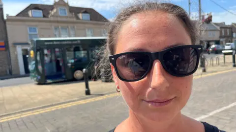 John Devine/BBC Ashley Whelton, 34, has brown hair pulled back tightly over her head, she is wearing dark glasses in strong sunshine on a street with a green coach at the bus stop behind her, shops and cars can also be seen in the background.