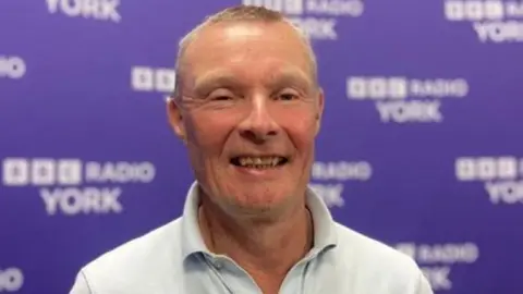 Ellis is positioned slightly off-center and is facing toward the camera. He is wearing a light blue, short-sleeved polo shirt with a small embroidered logo on the chest.
The background is a solid purple wall covered with repeating white text and logos. The text reads “BBC RADIO YORK” and is repeated multiple times in a regular pattern across the backdrop.