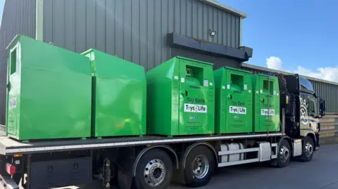 Toys 4 Life Five large shiny green recycling containers on the back of a lorry beside a warehouse on a sunny day