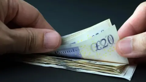 Getty Images A close up of a pair of hands, flicking through a pile of twenty pound notes against a black backdrop.