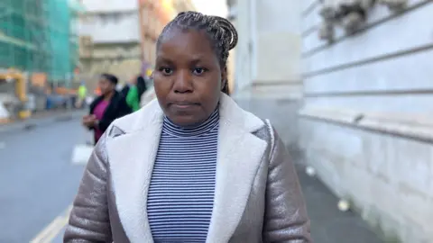 Nonkanyiso Tshuma standing outside Bristol Crown Court, wearing a black and white striped top and a metallic coat with a white fur lined collar. She has her hair in braids and tied back, and is looking at the camera with a forlorn expression. 