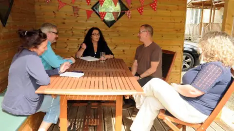 Five people are sitting in a covered wooden veranda in the Exhange's garden area. They are sitting around a wooden garden table. Some are on benches and some are on folding garden chairs. The Exchange building is just about visible in the corner of the photo and so is the front of a car. The three women and two men are in discussion. Two of the women have a pen in their hand and a notepad in front of them.