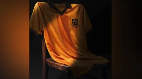 Old Gold Pack The orange and black shirt is draped on a chair. It has a V- neck in black with the friend's cross design on the left breast. 