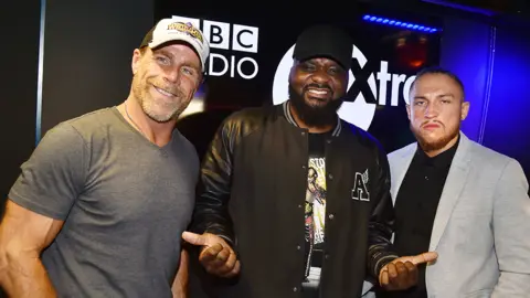 DJ Ace with WWE legend Shawn Michaels (left) who is wearing a grey tshirt, silver chain and cap and British wrestling star Pete Dunne (right) who is wearing a black top under a light grey blazer. Behind them is a black screen with BBC 1Xtra written in white.