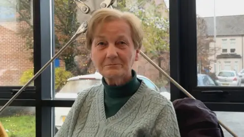 Eileen has blond hair and is wearing a dark green turtle neck jumper with a pale green v-neck jumper on top 