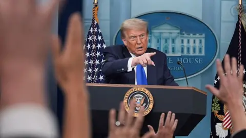Donald Trump at a podium pointing at the crowd of press