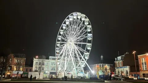 BBC A white ferris wheel with a crane behind it and buildings in the background