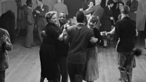 A black and white image shows people dancing in a club. They are smartly dressed and smiling