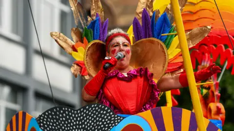 Pacemaker A person with red gloves and red hearts painted on their face is holding a microphone. They are wearing a multi-colour outfit and are sitting on top of a float.