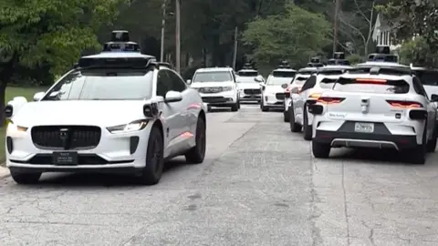 A litany of Waymo cars take up all the space in a street.