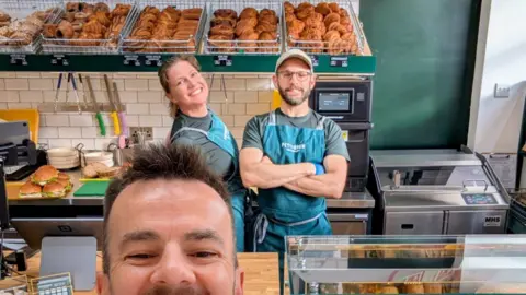 David Le Masurier David is only half pictured in this photograph. He is taking a selfie with his husband Lee and his colleague Alice in the background behind the counter. The three are smiling in a bakery with pastries in wire baskets on a shelf above their heads and filled bread rolls on a counter next to them.