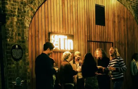 Robert Barrett Exterior of The Tin at the Coal Vaults independent music venue. A group of people are standing outside laughing with drinks. 