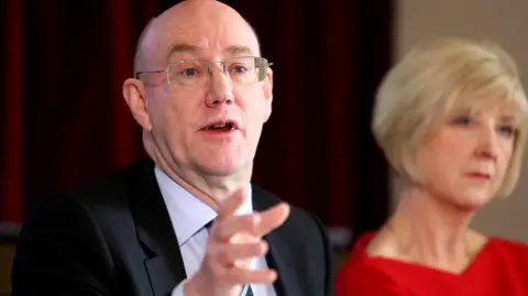 PA Media Dr Bill Kirkup answering questions at a press conference in 2015 as he presented the results of the investigation into the Morecambe Bay maternity scandal. He is a man in his 60s with clear glasses wearing a dark suit and white shirt.